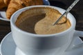 Thick foam of hot coffee cappuccino with spoon in white cup and croissant on wooden table, closeup. Milk foam cappuccino, latte Royalty Free Stock Photo