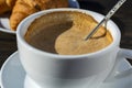 Thick foam of hot coffee cappuccino with spoon in white cup and croissant on wooden table, closeup. Milk foam cappuccino, latte Royalty Free Stock Photo
