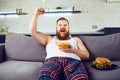 Thick funny man in pajamas eating a burger sitting on the couch.