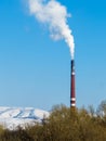 Thick white smoke comes out of the red pipe of the factory against a clear blue sky, snowy mountains and treetops.