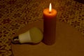 A thick yellow candle burns with a bright flame, and an LED light bulb lies next to it. Royalty Free Stock Photo