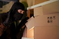 A thief wearing a ski mask picks up a box with a parcel from a warehouse at night