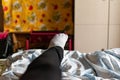 Thin female legs and feet in black pants and gray socks lie on the bed among colored fabric textiles