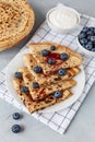Thin pancakes on plate on white checkered napkin with blueberries and sour cream. Grey background.