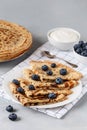 Thin pancakes on white plate with blueberries and sour cream. Served with a white checkered napkin and small teaspoon.