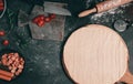 Thin pizza dough on a cutting board with a rolling pin, chopped cherry tomatoes and sausages on a dark background