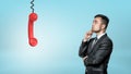 A thinking businessman looks up on a red retro phone receiver hanging from a black cord. Royalty Free Stock Photo