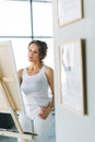 Thinking charming young woman artist in casual clothes with paint brushes in front of easel in modern art gallery Royalty Free Stock Photo