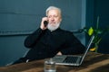Thinking gray-haired mature aged business man talking on mobile phone sitting at wooden table with laptop computer. Royalty Free Stock Photo
