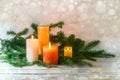 Third Advent, arrangement with four candles in orange and yellow, three are lit with a flame, fir branches on light rustic wood,