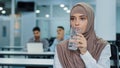 Thirsty millennial indian arabic female worker in hijab holding glass drinking pure mineral water in office, thoughtful Royalty Free Stock Photo