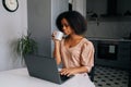 Thoughtful African American female freelancer typing on laptop looking to device screen and drinking coffee or tea in Royalty Free Stock Photo