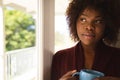 Thoughtful african american woman looking out of sunny window holding cup of coffee Royalty Free Stock Photo