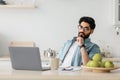 Thoughtful arab freelancer man looking at laptop screen while working remotely at home, sitting at kitchen with computer Royalty Free Stock Photo