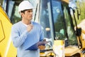 Thoughtful architect with clipboard and pen standing against earthmover at site Royalty Free Stock Photo