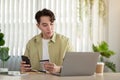 A thoughtful Asian man holding his smartphone and credit card and looking at his laptop screen Royalty Free Stock Photo