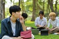 Thoughtful asian man student sitting on green grass in front of university building. Education, technology and lifestyle