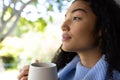 Thoughtful biracial woman holding cup of coffee and looking out window at home