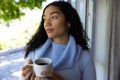 Thoughtful biracial woman holding cup of coffee and looking out window at home