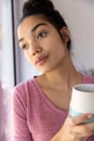 Thoughtful biracial woman holding cup of coffee and looking out window at sunny home Royalty Free Stock Photo