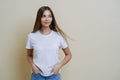 Thoughtful brunette woman looks aside, wears casual white t shirt, keeps hands in pockets, looks aside, isolated over beige Royalty Free Stock Photo