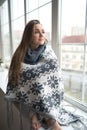 Thoughtful calm pensive young woman sitting on window sill at home wrapped in warm comfy sweater