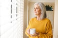 Thoughtful caucasian senior woman standing looking out of window at home with coffee, smiling Royalty Free Stock Photo