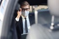 Thoughtful confident businessman in medical mask keeping hand on glasses while sitting in the luxe car