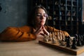 Thoughtful cute young woman in elegant eyeglasses thinking about chess move sitting on floor in dark library room Royalty Free Stock Photo