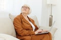 a thoughtful, enthusiastic elderly woman in a stylish brown suit sits on a beige sofa with a laptop on her lap and looks
