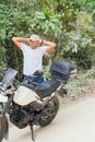 Thoughtful ethnic young man in casual clothes and cap standing near a modern motorcycle with his hands behind his head and looking