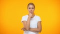 Thoughtful female with hand on chin standing on orange background, hesitation