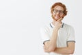 Thoughtful handsome young genious in glasses with red hair standing in thinker pose with hand on chin gazing with smart