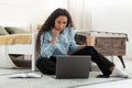 Thoughtful millennial female using laptop pc, sitting on floor with coffee at home, full length