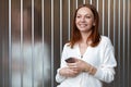 Thoughtful pleased woman with dreamy look, holds mobile phone, sends text message, wears white formal jumper, connected to