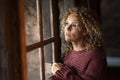 Thoughtful portrait of attractive adult woman at home looking outside the windows reflected on the gass. Worried adult female