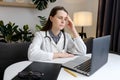 Thoughtful sad young woman doctor sit at work desk with pc laptop, pondering problem solution, pensive therapist physician wearing