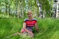 The thoughtful woman of average years sits on a grass in the birch wood