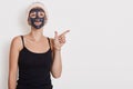 Thoughtful woman looks pensively up, applies black mask on face, wears hairband and sleeveless t shirt, points away at empty space Royalty Free Stock Photo