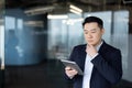 A thoughtful young Asian man is standing inside an office and looking seriously at the screen of a tablet he is holding in his Royalty Free Stock Photo