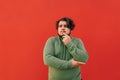 Thoughtful young bearded corpulent hispanic man is standing on a red background, expressing a thinking gesture, touching his chin Royalty Free Stock Photo