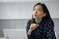 Thoughtful young business Asian woman holds a pen and looks out the window while working with a laptop Royalty Free Stock Photo