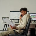 Thoughtful young male trader holding papers with statistics while talking on the phone, sitting at desk, trading from