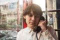 Thoughtful young man in light striped shirt makes call from old public telephone booth, city reflections in background Royalty Free Stock Photo