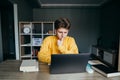 Thoughtful young man in a yellow sweatshirt sits at home at a table in the room and uses a laptop, pensively looking at the screen Royalty Free Stock Photo