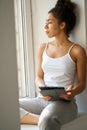 Thoughtful young woman with tablet pc looking out the window, feeling cozy while sitting alone by the window at home Royalty Free Stock Photo