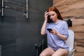 Thoughtful young woman using mobile phone sitting on toilet at bathroom, pensive looking to smartphone screen. Redhead Royalty Free Stock Photo