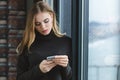 Thougtful woman standing near the window and using her smartphone