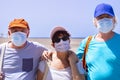 Three adult senior friends standing on the beach, group of people in outdoor vacation at sea wearing face mask due to coronavirus Royalty Free Stock Photo