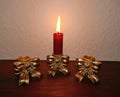 .Three antique golden candlesticks decorated with bows on a mahogany shelf in front of a bright wall. In one is a burning red cand